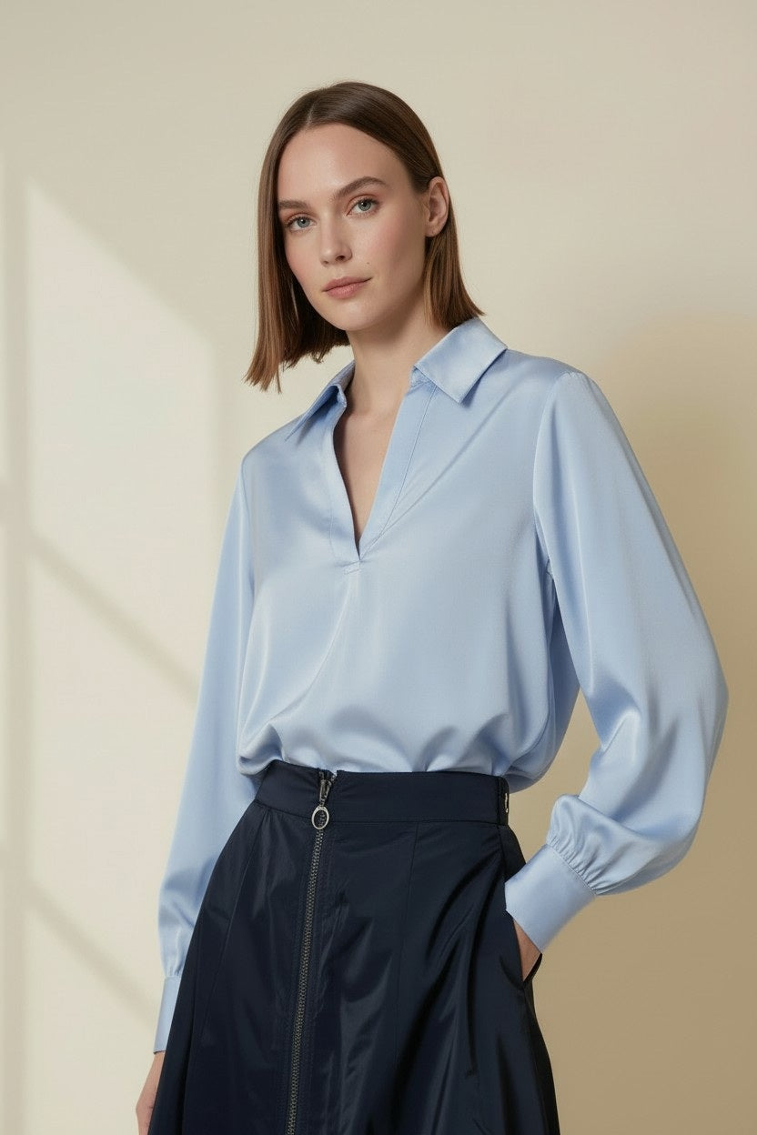 A woman with a bob hairstyle stands against a light beige background, wearing the VICARIO CINQUE Solei Taffeta Top in Light Blue—a relaxed fit, long-sleeved blouse—paired with a dark, high-waisted zippered skirt.