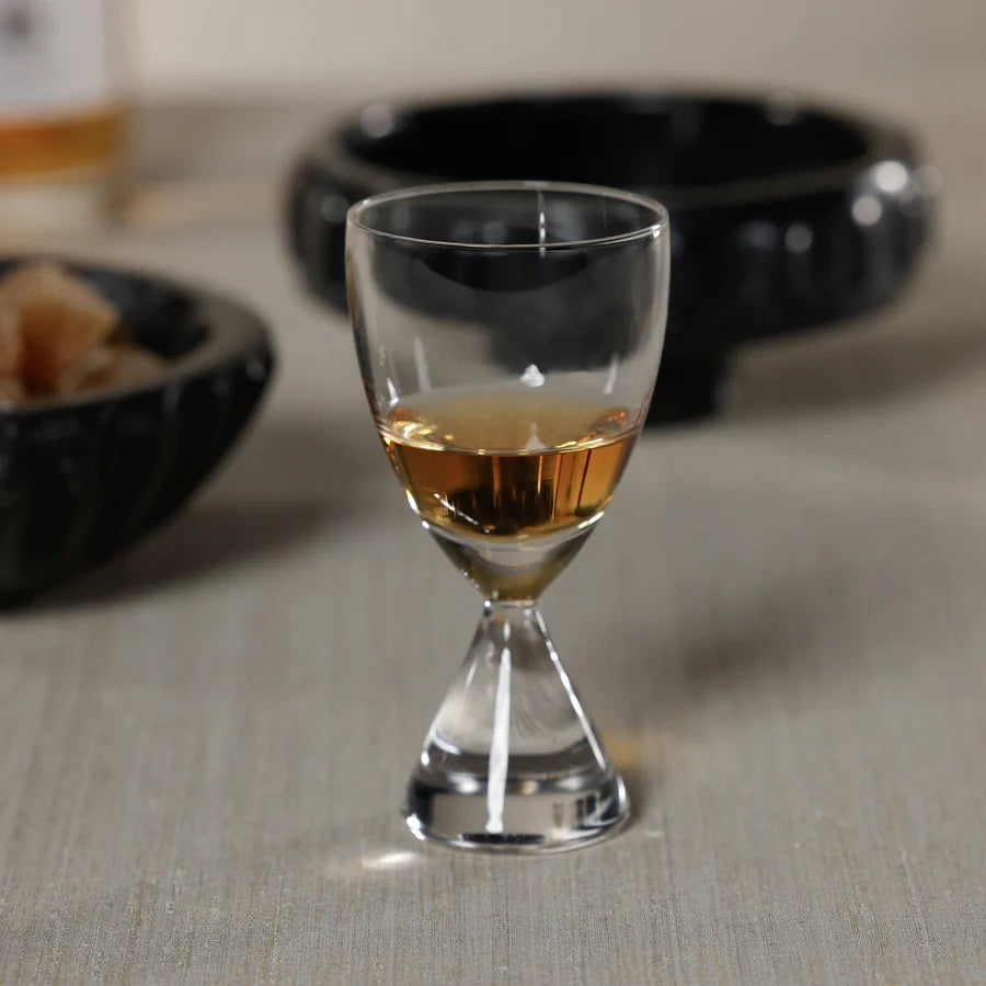 The ZODAX TIMELESS SIP SHOT GLASS, featuring a unique double-ended shape, sits half-filled with amber liquid on a light surface, while two dark round dishes are softly blurred in the background.