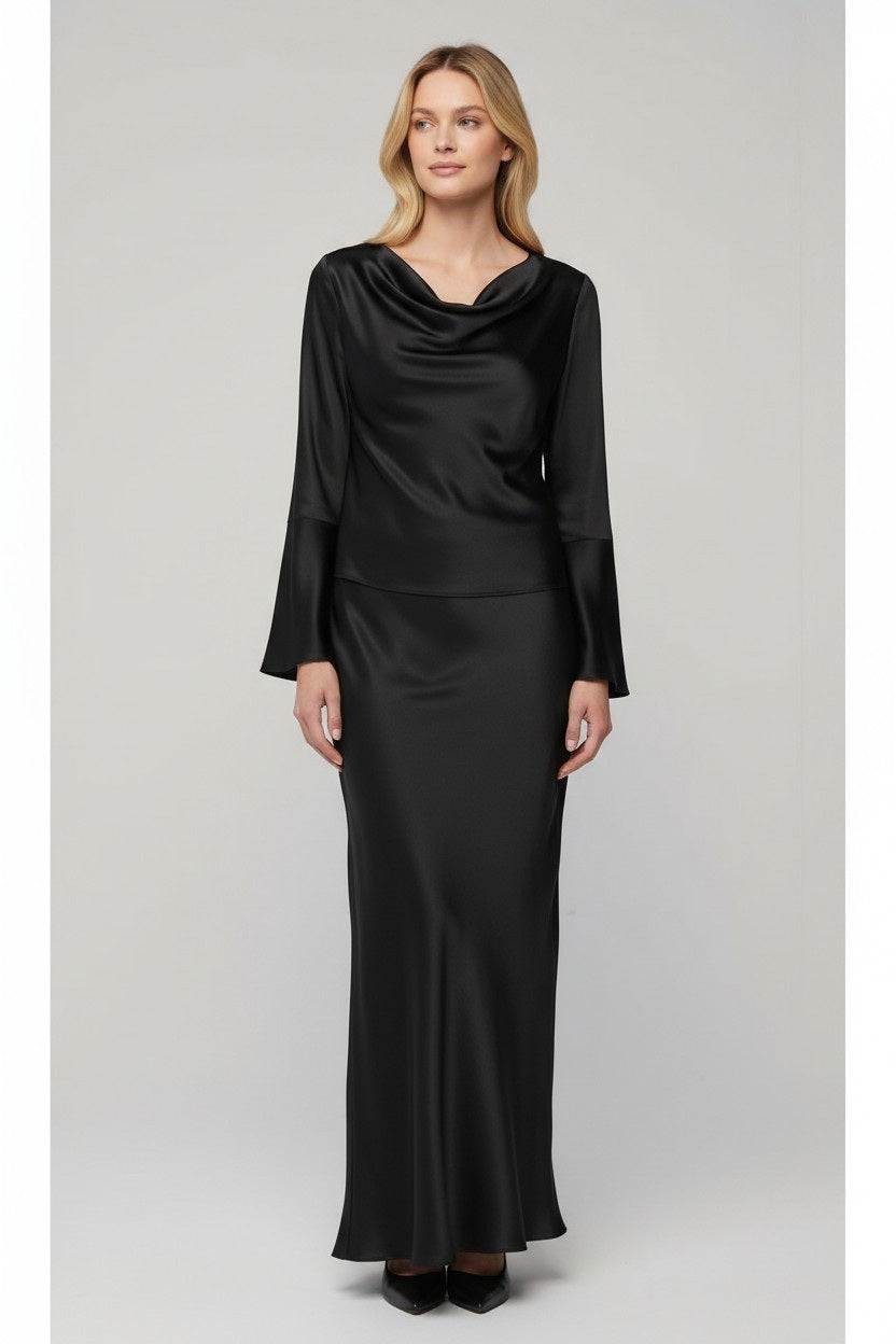 A woman stands against a light gray background, wearing the MILVA MI Satin Blouse in Black by MILVA. The blouse features long sleeves and a draped cowl neckline, and she pairs it effortlessly with wavy brown hair and a slight smile, exuding relaxed sophistication with its elegant silhouette.