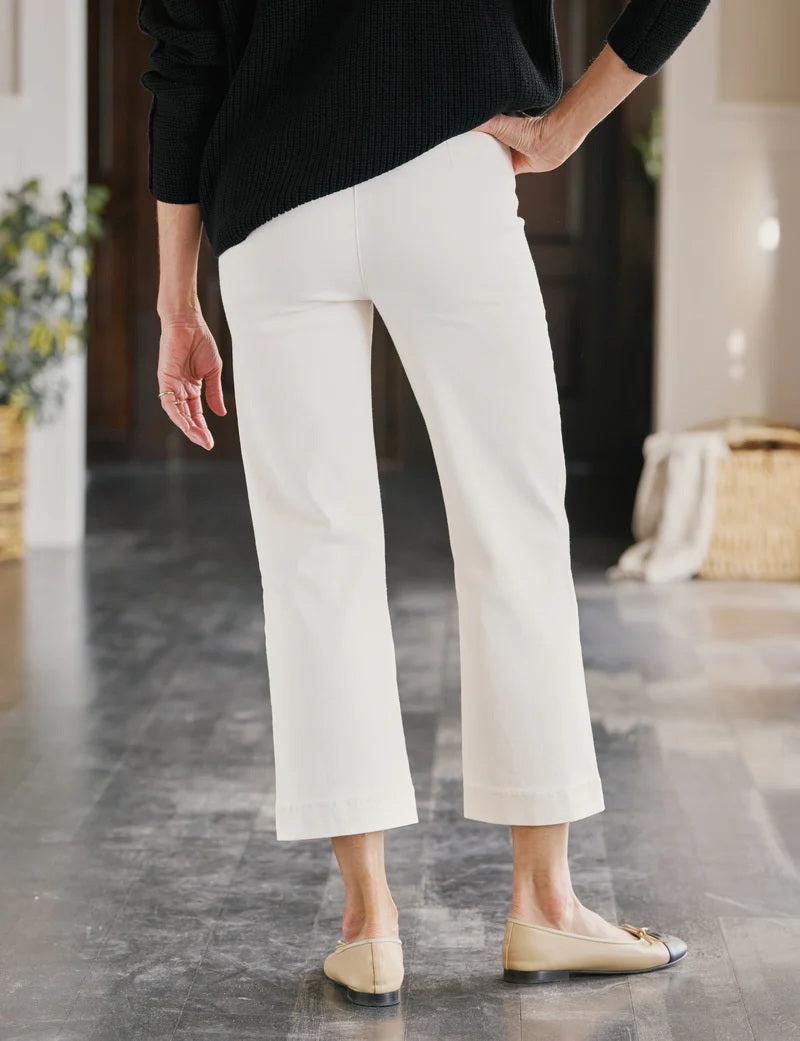 Wearing FRANK & EILEEN’s WESTPORT ILLUSION PULL ON WIDE LEG JEAN in Antique White, a person pairs them with a black sweater and beige flats with black toes, standing on a dark wooden floor in a bright, modern room.