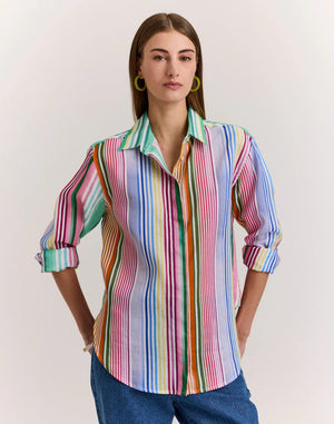 A woman in the HINSON WU Halsey Long Sleeve Linen Shirt in Multi Stripe, featuring vibrant vertical stripes, rolled-up sleeves, and lightweight linen fabric, pairs it with blue jeans. She has straight brown hair, hoop earrings, and a bracelet.