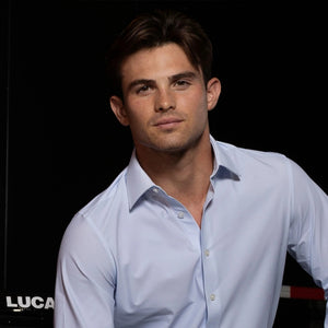 A young man with short brown hair poses against a dark background, wearing the COLLARS AND CO - APEX DRESS SHIRT SEMI SPREAD IN GLACIER BLUE TWILL by COLLARS AND CO. He gazes at the camera with a neutral expression. The partial word “LUCAS” appears to his left, evoking a sense of excellence reminiscent of the Arrow McLaren IndyCar Team.