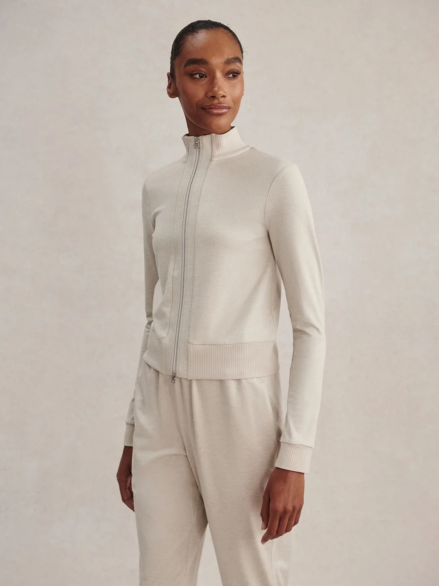 A person wears a light beige Varley Rufus Cropped Zip Through Sweat in Pebble Marl with a high collar and matching pants.
