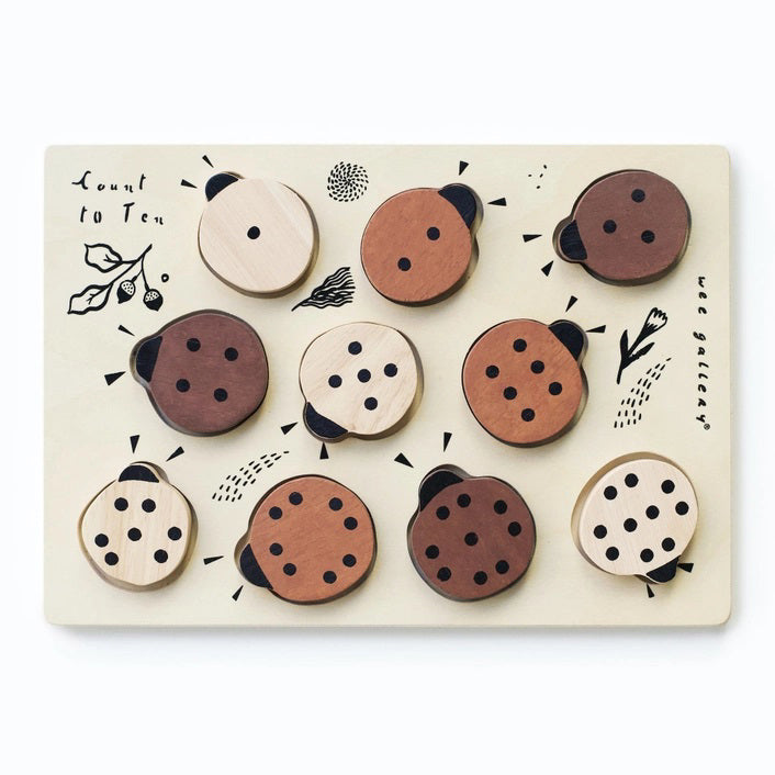 The WEE GALLERY - LADYBUGS COUNT TO 10 TRAY PUZZLE by WEE GALLERY is a wooden puzzle board featuring ladybug-shaped pieces in varying shades from light beige to dark brown. This educational puzzle helps teach counting skills and depicts leaves and twigs alongside the words "Count to Ten" on the left side. Each ladybug is adorned with black dots on their backs to represent number concepts.