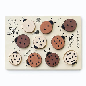 The WEE GALLERY - LADYBUGS COUNT TO 10 TRAY PUZZLE by WEE GALLERY is a wooden puzzle board featuring ladybug-shaped pieces in varying shades from light beige to dark brown. This educational puzzle helps teach counting skills and depicts leaves and twigs alongside the words "Count to Ten" on the left side. Each ladybug is adorned with black dots on their backs to represent number concepts.