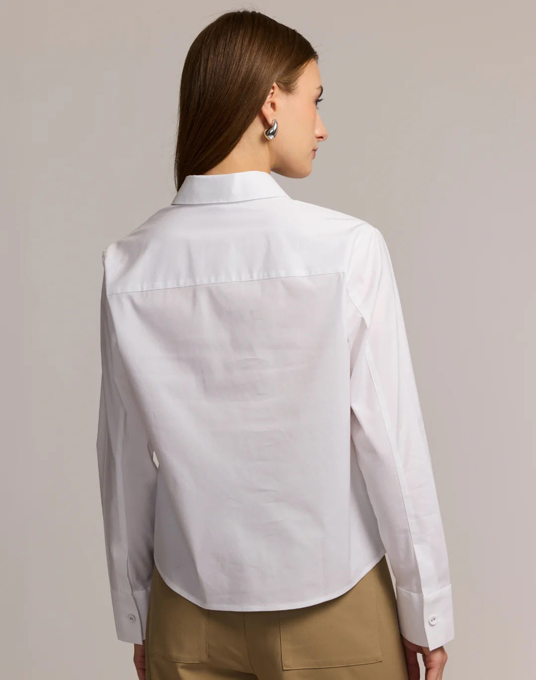 A woman models the HINSON WU - VENUS LONG SLEEVE UNTUCKABLE SHIRT in white stretch polished cotton with a chest pocket, paired with beige pants, standing against a neutral background and holding the shirt collar.