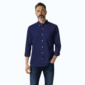 A man with short dark hair and a beard stands sideways with arms crossed, wearing the FRANK & EILEEN LUKE Men's Button Up Shirt in Famous Denim Washed Navy, blue jeans, and white sneakers. He is indoors by a light wall.