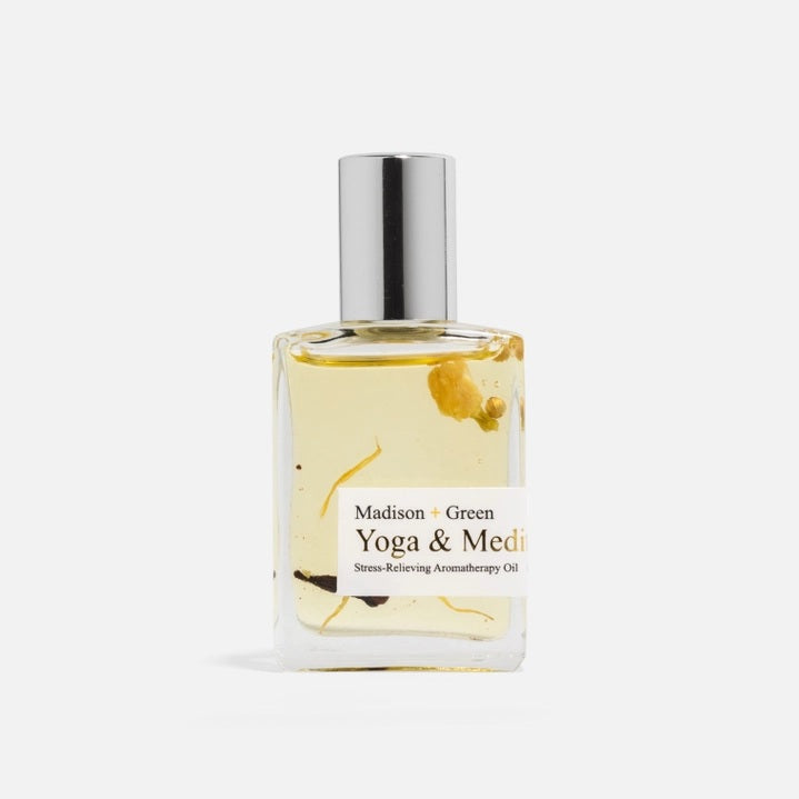 A clear glass bottle with a silver cap holds yellow aromatherapy oil labeled “MADISON AND GREEN - YOGA & MEDITATION SPIRITUAL AROMATHERAPY BODY OIL MINI” from MADISON GREEN, with visible herbs or botanicals suspended inside—making this botanical body oil ideal for self-care rituals.