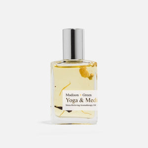 A clear glass bottle with a silver cap holds yellow aromatherapy oil labeled “MADISON AND GREEN - YOGA & MEDITATION SPIRITUAL AROMATHERAPY BODY OIL MINI” from MADISON GREEN, with visible herbs or botanicals suspended inside—making this botanical body oil ideal for self-care rituals.