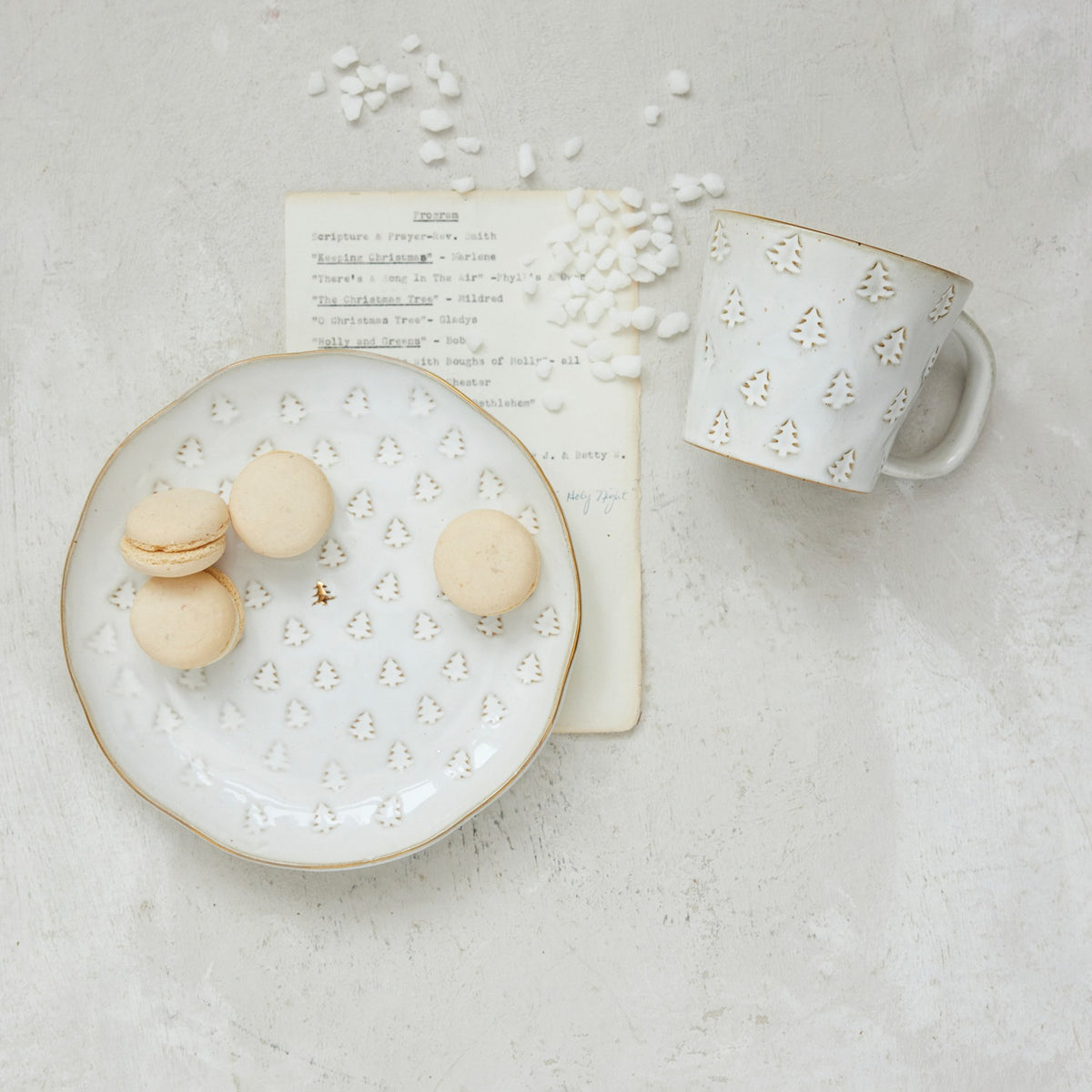 A DEBOSSED PLATE WITH GOLD ELECTROPLATING and matching mug with a reactive glaze and debossed gold Christmas tree patterns, containing macarons and marshmallows next to a printed holiday program.