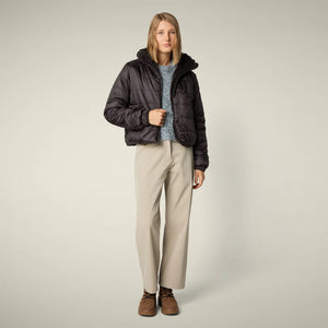 A woman models the SAVE THE DUCK - ELINA REVERSIBLE JACKET in beige fur, styled with a white skirt, orange tights, burgundy boots, and a matching clutch, standing against a plain light background.