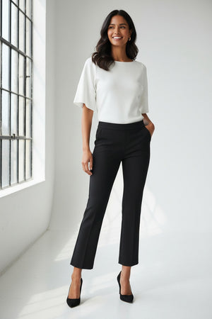 A woman stands confidently with hands on hips, wearing a white short-sleeve top, black SPANX - PERFECT PANT KICK FLARE from SPANX, and black heels against a plain light background.
