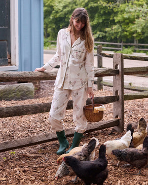 A woman dressed in the PRINTFRESH Embroidered Hens Long Pajama Set in Buff smiles as she stands by a wooden fence, holding a basket and watching chickens peck at the ground in a farmyard setting, with trees and a path visible behind her.
