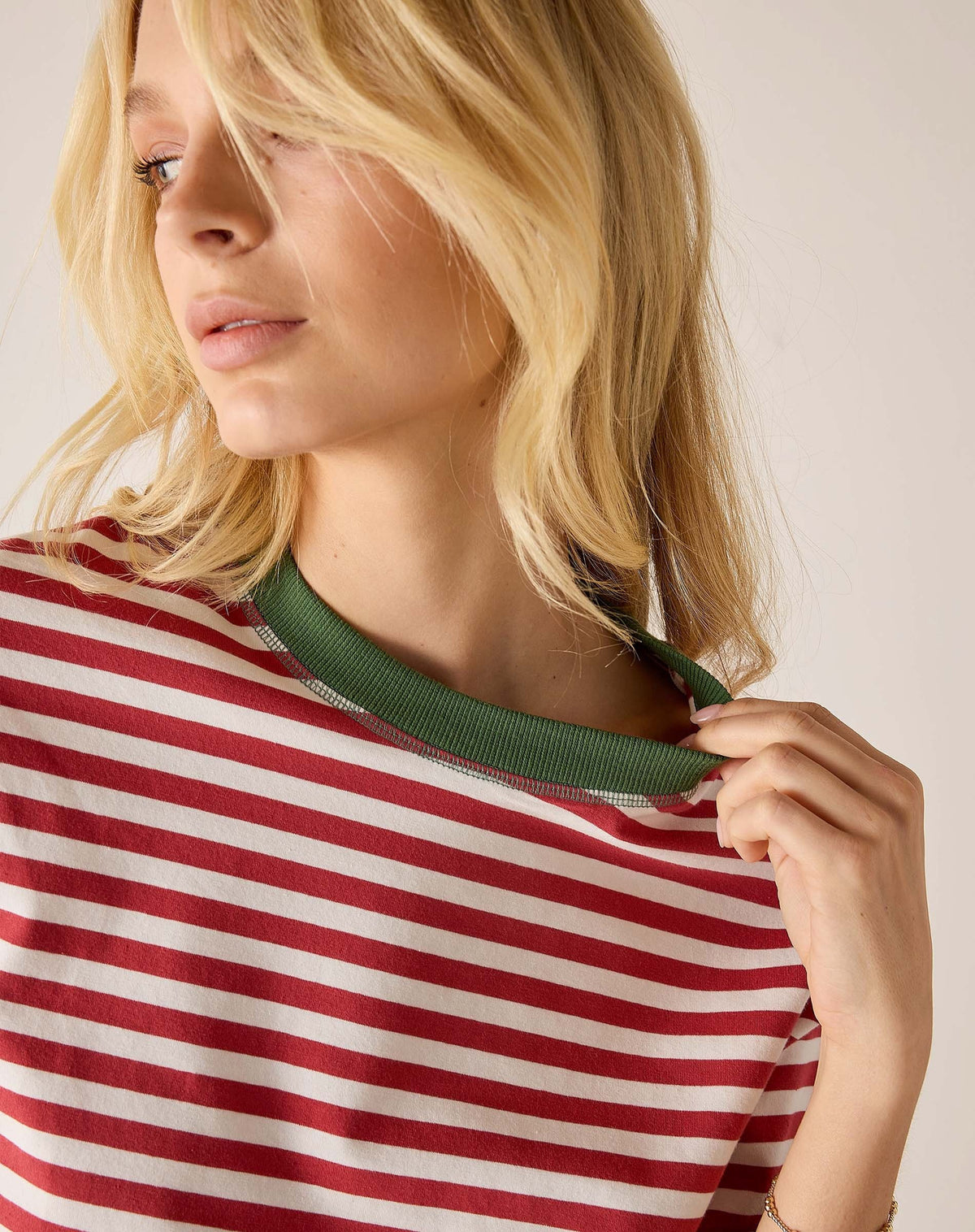 A smiling person with long blonde hair wears the MERSEA Deauville Ringer Tee in Currant Jade—a red and white striped ringer tee with green collar and cuffs—paired with green pants, standing against a light background.