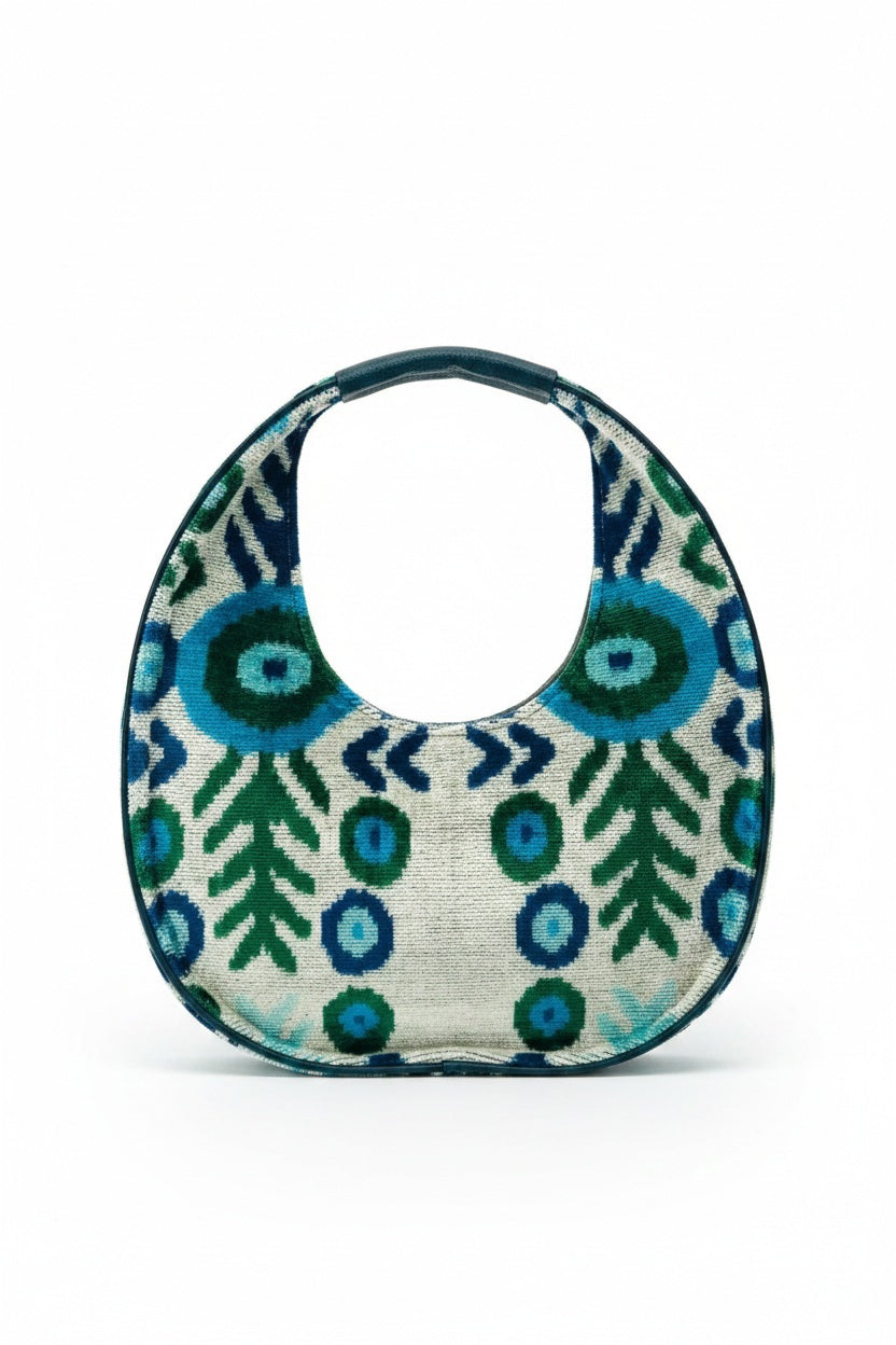The BALDIZ LUCA BAG IN AQUA VINE is a circular handbag with a short handle, showcasing bold blue and green abstract shapes and leaves on a light background—perfect for everyday use and inspired by the signature style of BALDIZ.