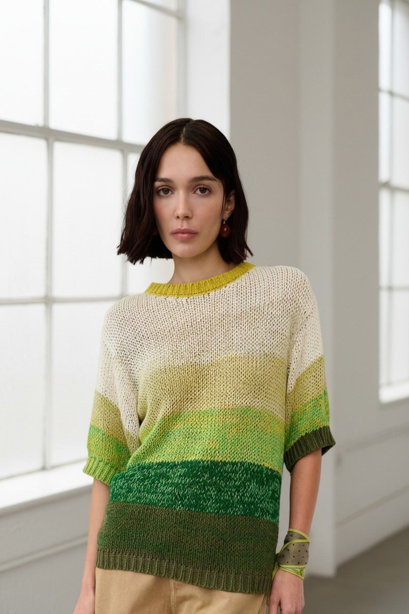 A woman with shoulder-length dark hair wears the FLOOR - Cotton Sweater in Thyme, featuring green and cream stripes, paired with beige pants as she stands in a bright room with large windows.