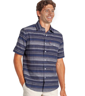 A man smiles with one hand in his pocket, wearing the CRIQUET Hemp Stripe Pearl Snap Shirt in Navy—short-sleeve, button-up with white stripes—paired with beige pants.