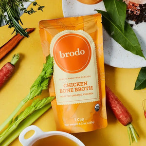 A pouch of BRODO BROTH COMPANY’s BRODO BROTH- CHICKEN BONE BROTH ORGANIC rests on a yellow surface, surrounded by carrots, herbs, and a bowl of collagen-rich broth. The orange packaging with white accents is labeled “Roasted Organic Chicken,” showcasing this keto friendly chicken bone broth as a nourishing addition to any meal.