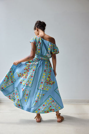 A woman with short curly hair sits on a rustic green bench, wearing the HOPE FOR FLOWERS - Flared Dress in Chambray Floral by HOPE FOR FLOWERS and brown sandals, looking thoughtfully to the side with her hands in her lap.
