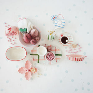 A flat lay of HANDPAINTED STRIPED PLATE WITH TREE, assorted stoneware plates, bowls with red and green stripes, a holly leaf platter, pink candies, paper trees, and a tea set on a white polka-dot surface.