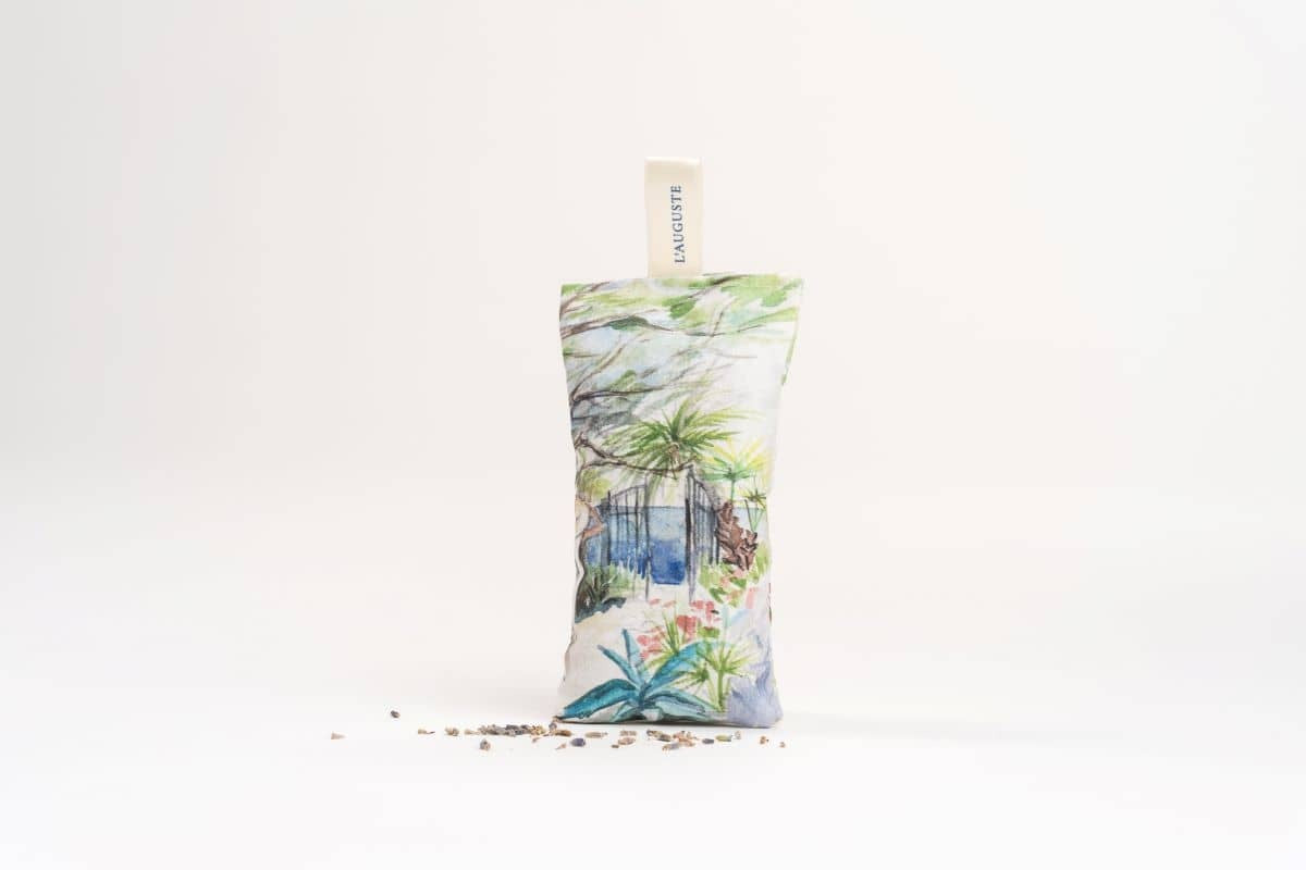 A L'AUGUSTE PROVENCE lavender sachet with a tropical botanical print stands upright on a white background, surrounded by scattered seeds. The label “L'AUGUSTE PROVENCE” is visible at the top.