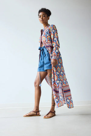 In a minimal room, a person with short curly hair and brown sandals exudes confidence in the HOPE FOR FLOWERS BAND COLLAR SHIRTDRESS IN CHAMBRAY MEDALLION, showcasing modern style from the HOPE FOR FLOWERS brand.