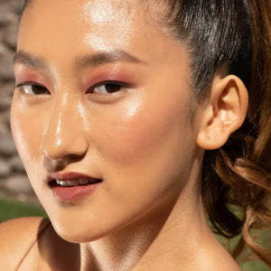 A close-up of a person with curly dark hair, a serious expression, and warm makeup featuring the universally flattering, weightless JASON WU - HOT FLUFF LIP/EYE/CHEEK by JASON WU BEAUTY on their face. They are looking directly at the camera with a neutral background.