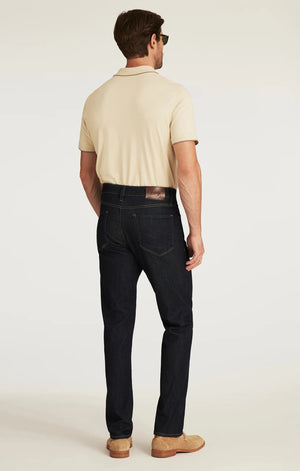 A man wearing a beige short-sleeve shirt, tan suede shoes, and the 34 HERITAGE - CHARISMA RELAXED STRAIGHT JEANS IN RINSE VINTAGE by 34 HERITAGE stands with one hand in his pocket against a white background.