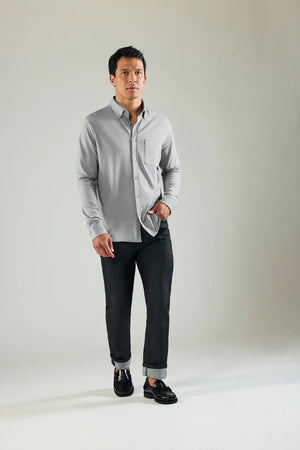 A man faces forward against a plain background, buttoning up a Monument-colored MACK WELDON Silver Pique Long Sleeve Button Up Shirt. He looks slightly upward with a neutral expression and pairs the shirt with dark pants.