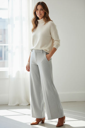 Indoors, a person wears FRANK & EILEEN's RILEY Wide Leg Trouser Sweatpant in light gray with a button waistband and a white long-sleeve top, one hand in their pocket. A red desk, wooden floor, and white cabinets complete the California Heritage Collection scene.