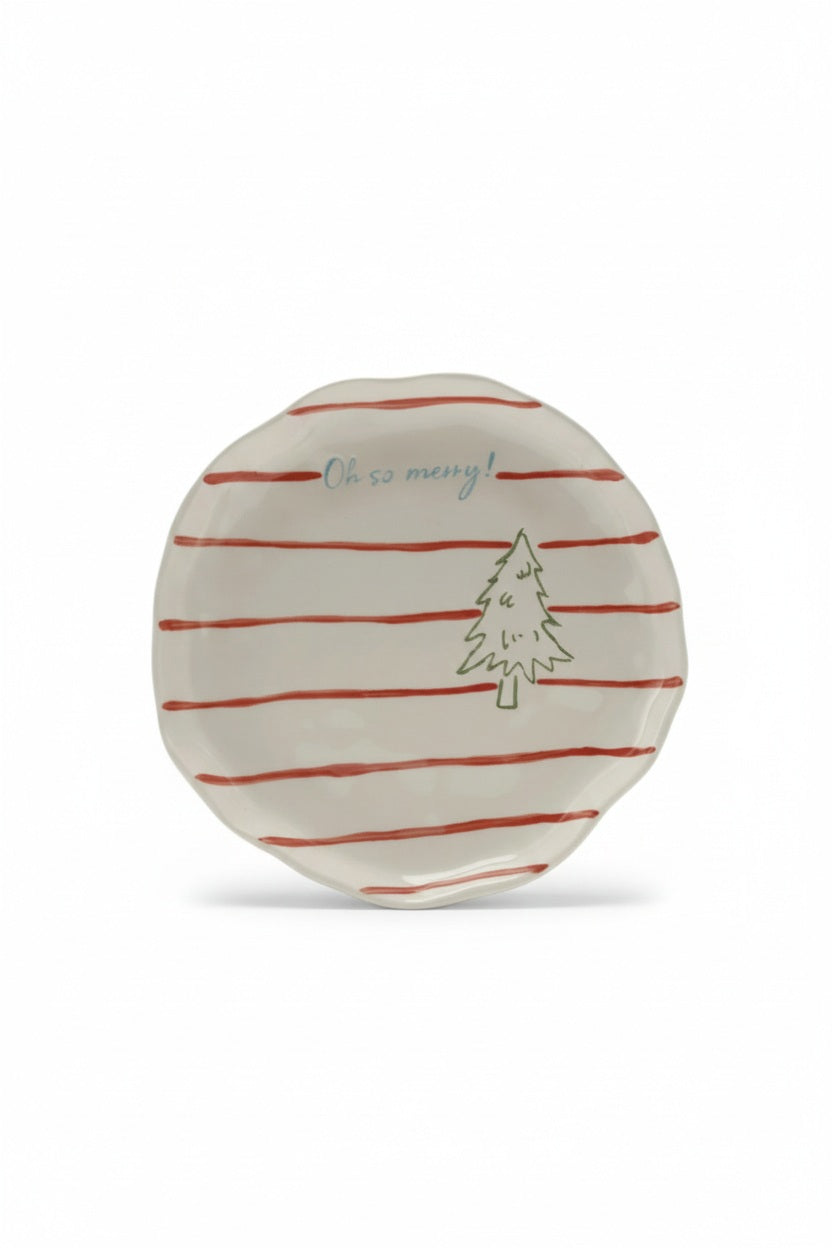 A flat lay of HANDPAINTED STRIPED PLATE WITH TREE, assorted stoneware plates, bowls with red and green stripes, a holly leaf platter, pink candies, paper trees, and a tea set on a white polka-dot surface.