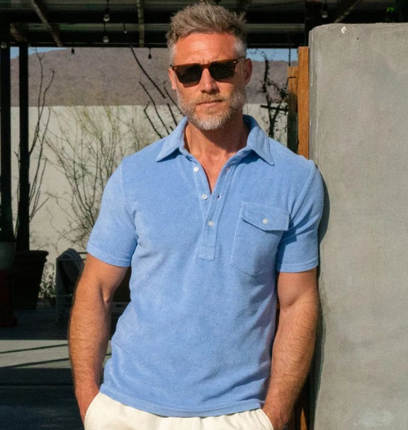 A person in a periwinkle Criquet Terrycloth Players Shirt and white shorts stands facing away from the camera against a plain beige background.