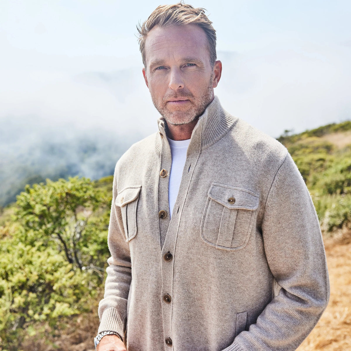 A man wearing the COLLARS AND CO Napoli Cardigan in Wool and Cashmere Camel stands outdoors among green shrubs and fog in a hilly landscape, looking to his left.