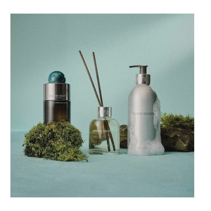 A bottle of MOLTON BROWN - COASTAL CYPRESS EAU DE PARFUM features an ocean-inspired scent, a gradient dark glass base, silver cap, and round blue-green marble-like top, displayed against a white background.