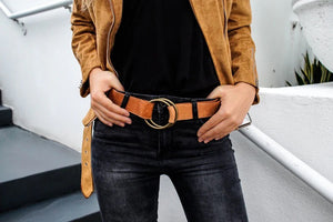 A person wearing a tan suede jacket, black shirt, and dark jeans stands on stairs, showcasing a tan belt with a large round gold buckle. Their hands rest on their hips.