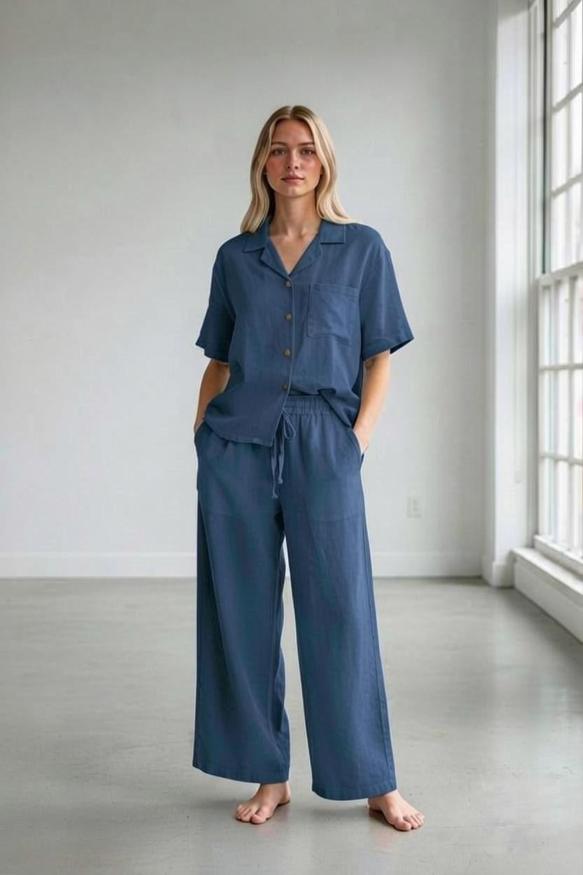 A woman stands barefoot in a bright room wearing a loose-fitting blue pajama set with short sleeves and wide-leg pants. She has long, blonde hair, and her hands are in the pockets of her LLEA BLUE - Danen Straight Leg Drawstring Linen Pant in Midnight Ocean.