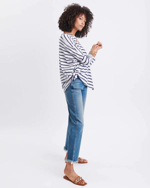 A woman with wavy brown hair models the MERSEA - CATALINA SLUB TEE—an oversized, long-sleeved white top with thin gray stripes—by MERSEA. She pairs it with blue jeans and stands against a plain, light background, hands behind her back.