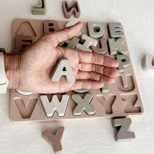 The ABC SOFT SILICONE PUZZLE from ALI + OLI sits on a light-colored surface, featuring cut-out slots for the entire alphabet. Some letter pieces are placed in their corresponding slots, while others are scattered around the board. This educational toy, with letters in various shades of beige, gray, and brown, promotes hand-eye coordination.