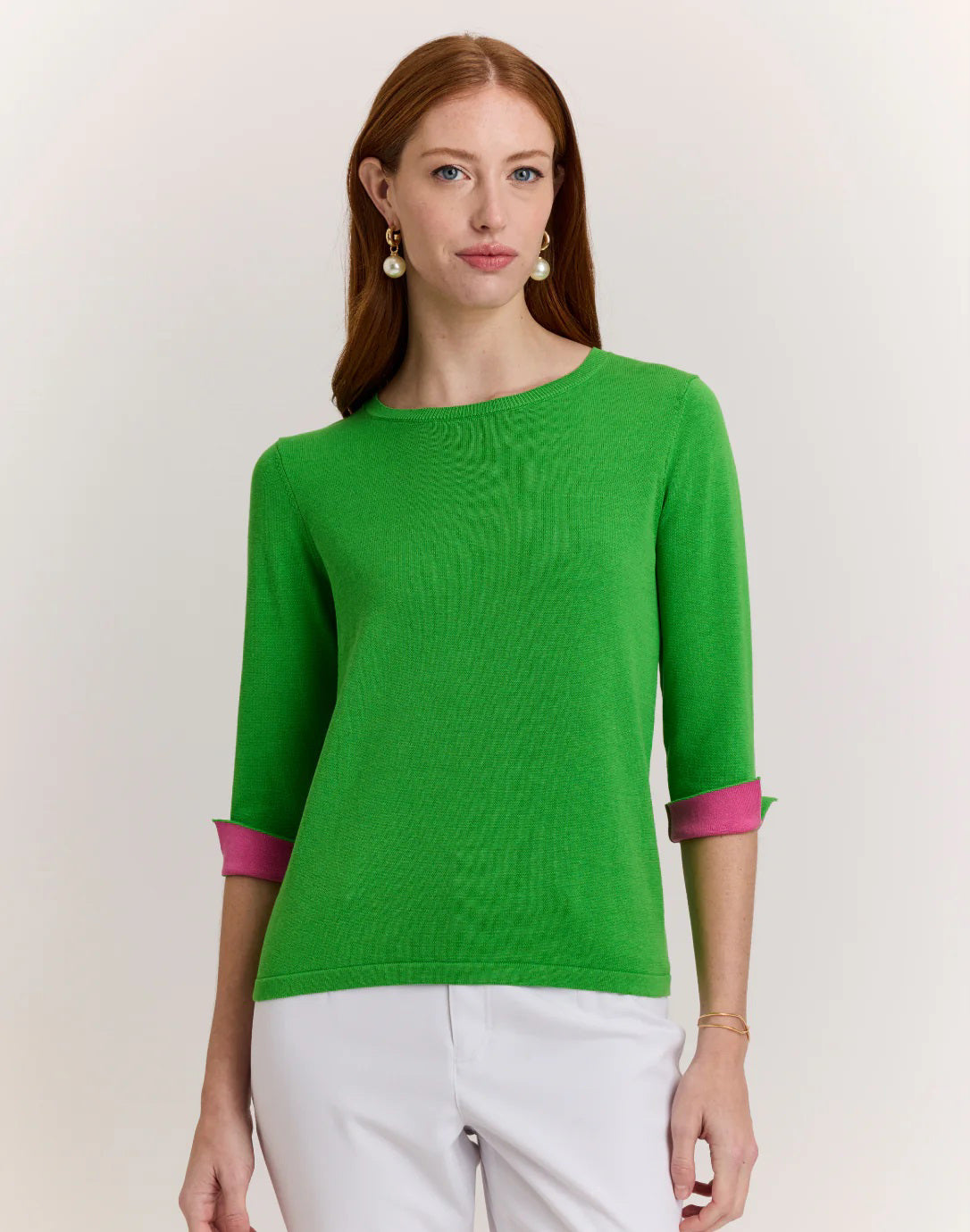 A woman with long red hair wears the HINSON WU TYLER 3/4 SLEEVE KNIT SWEATER in bright green with pink cuffs and white pants, standing against a plain light background and looking at the camera with a neutral expression.
