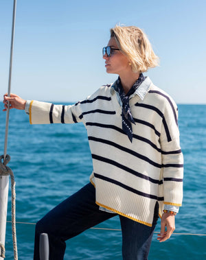 Wearing MERSEA’s Montauk Colorblock V Neck Sweater in Cream Stripe, a person with sunglasses and a scarf stands on a boat, holding the railing against a backdrop of calm blue water and clear sky.