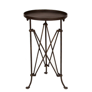 The CREATIVE COOP BRONZE FINISH METAL ACCENT TABLE holds a glass jar candle and a small flowering plant on a wooden floor, with framed artwork and photos displayed on the light-colored wall behind it.