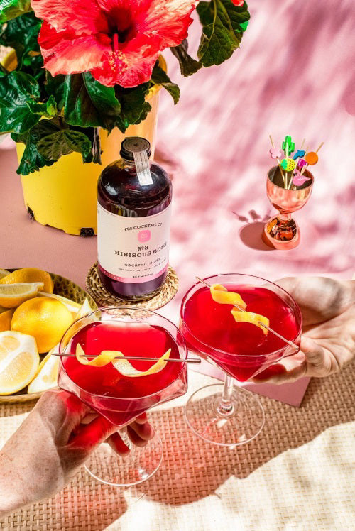 A transparent glass bottle holds a rich, dark red liquid labeled "YES COCKTAIL CO - Hibiscus Rose Cocktail Mixer." Handcrafted in California and available in a 16 fl oz (473ml) size, the white-and-red-toned label indicates its perfect suitability for creating cocktails or an exotic floral spritzer.