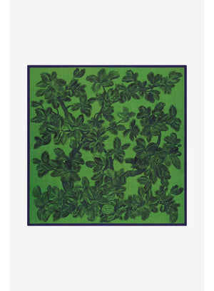 A person wearing a cream-colored suit and the INOUI EDITIONS - ARNORIS WOOL SCARF IN GREEN by INOUI, elegantly draped like a 100% wool shawl made in India, stands facing forward against a plain light background.