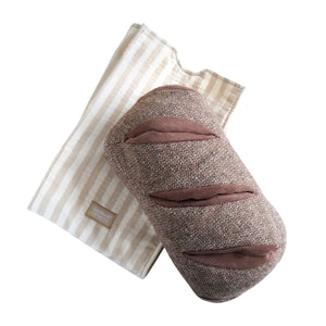 A fluffy brown dog is lounging on a soft, light-colored couch with its front paws resting on a brown pillow and its favorite LAMBWOLF COLLECTIVE - RYE DOG TOY nearby. The dog looks relaxed and content in a cozy, neutral-toned setting.