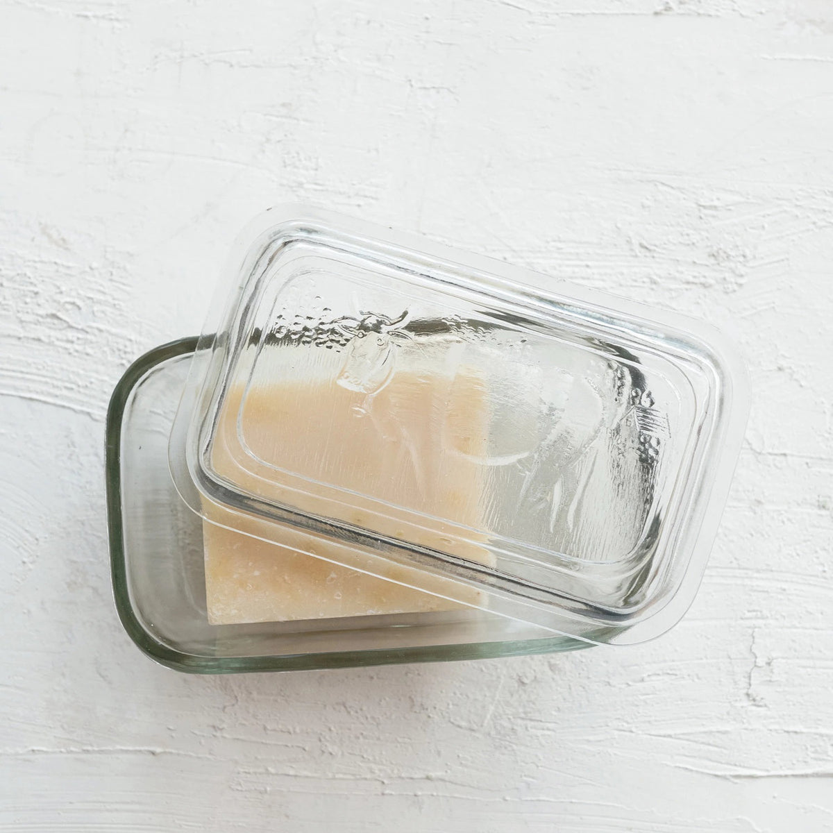 The PRESSED GLASS BUTTER DISH WITH COW by CREATIVE COOP holds a block of hard cheese. This rectangular, embossed glass dish with a transparent lid is placed on a light-colored, textured surface in a rustic kitchen, with the cheese partially visible through the lid.