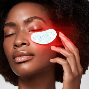 A crescent-shaped Omnilux Eye Brightener Kit is shown against a light gray background, featuring a power button and branding text. The device has a leaf pattern and emits a subtle red glow, highlighting its red light therapy benefits for collagen production under the eyes.