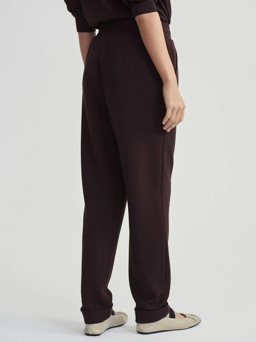 A person seen from behind wearing VARLEY - ROLLED CUFF PANT 25” IN COFFEE BEAN dark brown trousers with rolled cuffs and cream-colored ballet flats.