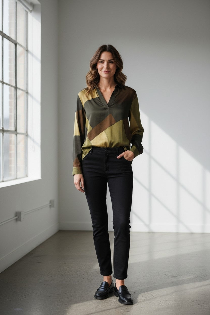 A woman stands in a bright room, smiling. Radiating French elegance, she wears the HARTFORD - CHORUS POPOVER SHIRT IN GREEN by HARTFORD, paired with blue jeans and tan loafers, her hands tucked into her jean pockets. Sunlight pours through large windows beside her.