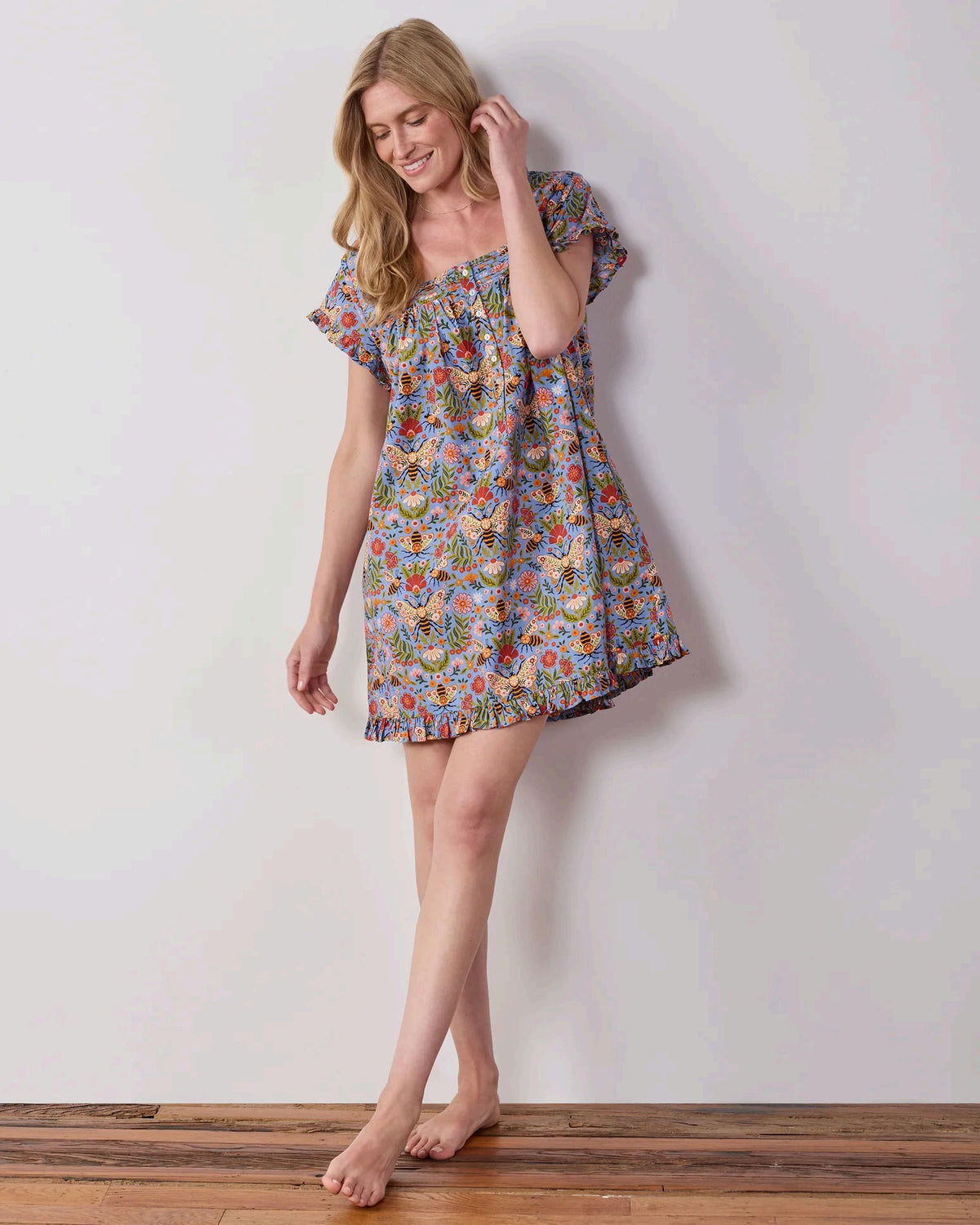A woman with long blonde hair wears the PRINTFRESH Bee Tapestry Pinktuck Nightgown in Delphinium Blue, standing against a plain light-colored wall and smiling slightly.