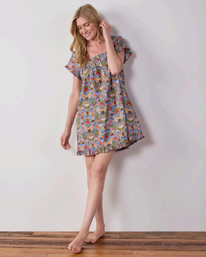 A woman with long blonde hair wears the PRINTFRESH Bee Tapestry Pinktuck Nightgown in Delphinium Blue, standing against a plain light-colored wall and smiling slightly.
